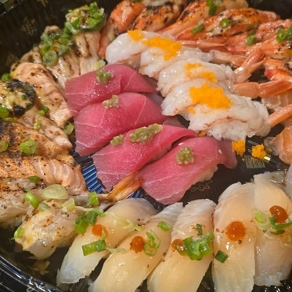 Assorted sushi and sashimi, including tuna, shrimp, salmon, and white fish, arranged in black takeout trays with dipping sauces.