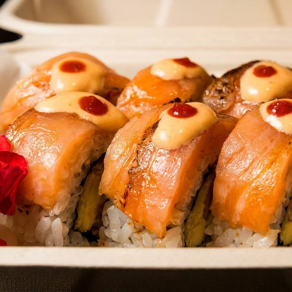 Sushi rolls with salmon, creamy sauce, and red dots, garnished with a red flower in a takeout container.