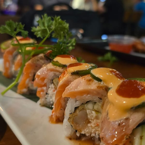Salmon-topped sushi rolls with creamy sauce and cucumber garnish on a white plate in a dimly lit restaurant.