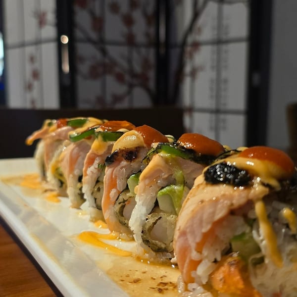 Close-up of salmon-topped sushi rolls with avocado, cucumber, sauce, and sesame on a white platter.